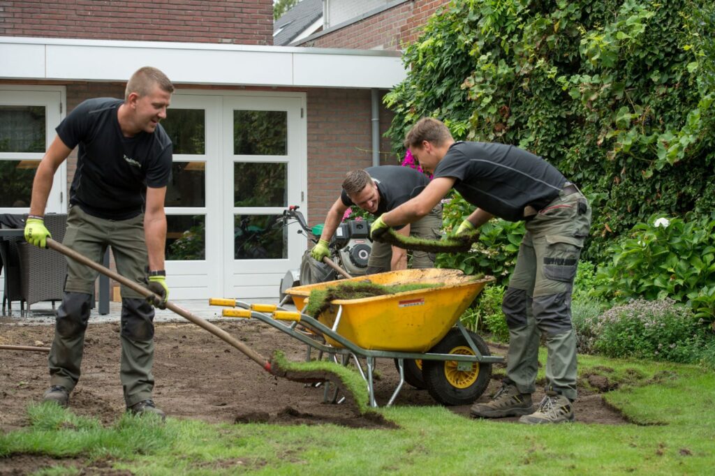 3 Männer arbeiten im Garten. Ein Mann schaufelt mit einer Schaufel Torf in eine Schubkarre.
